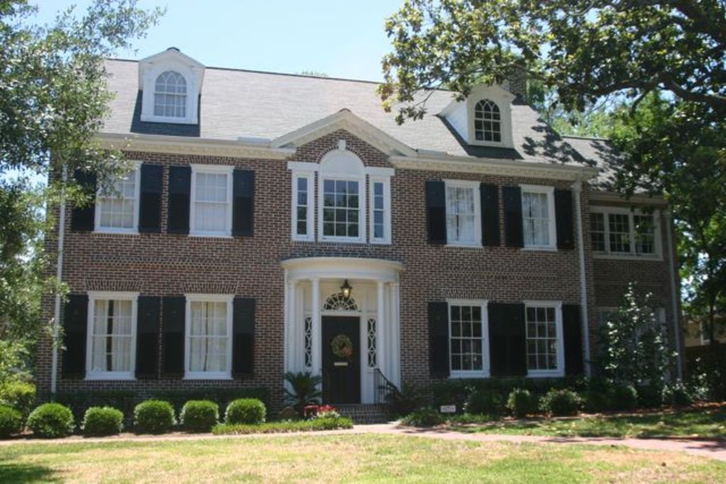 A large brick colonial home with dark shutters behind a neat front garden